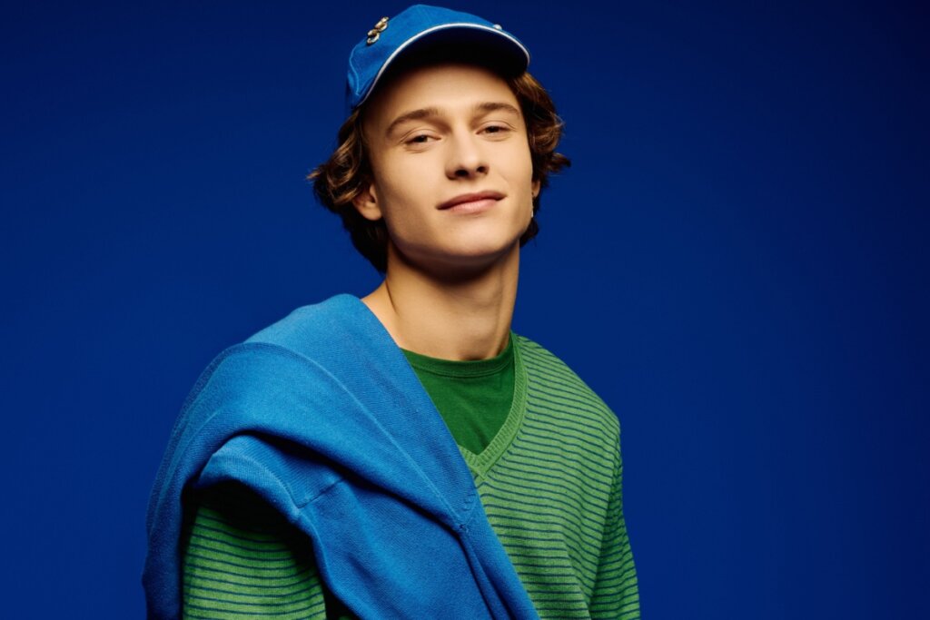 Homem sorrindo, usando boné azul, blusa de frio verde e casaco azul nos ombros, em frente a um fundo azul