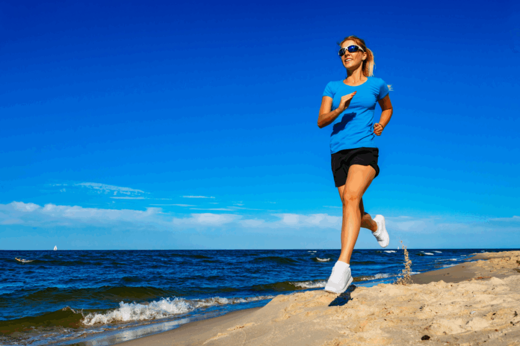 Mulher, usando blusa azul, short preto e óculos de sol, correndo na praia