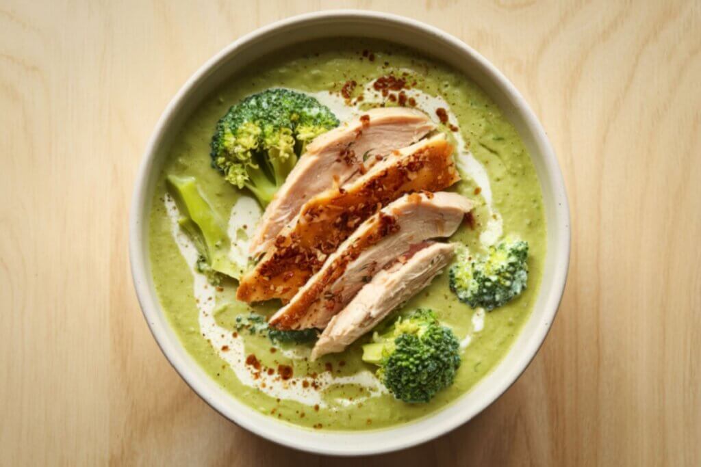 A imagem mostra uma tigela com um creme verde espesso, aparentemente feito à base de vegetais, acompanhado por pedaços de frango fatiado e floretes de brócolis.