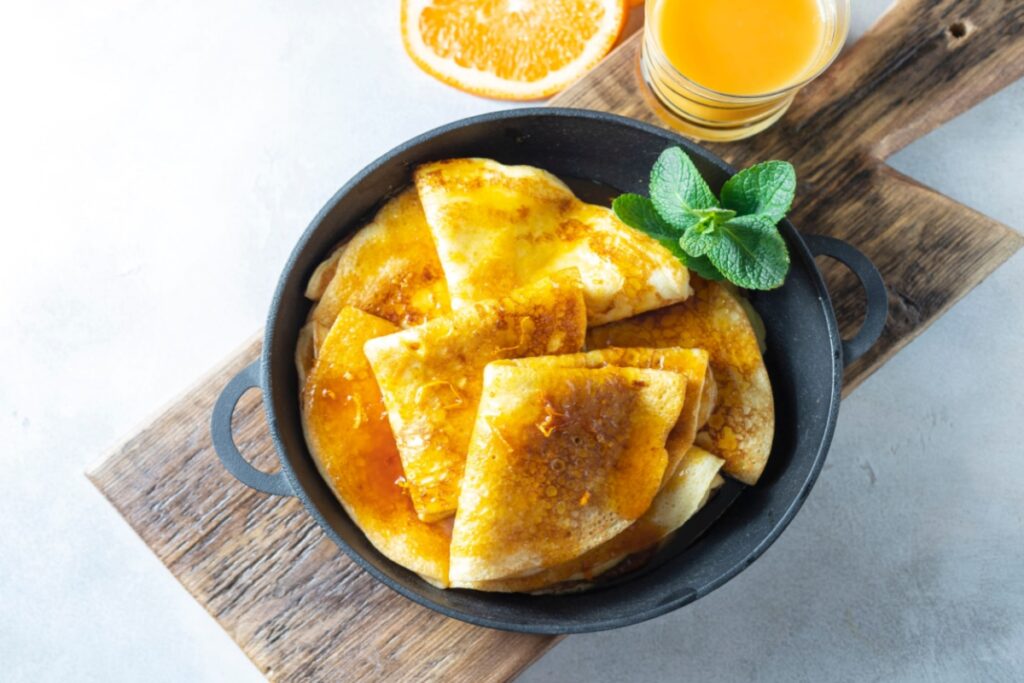 A imagem mostra um prato de crêpes dobrados e cobertos com uma calda alaranjada brilhante, acompanhados por uma fatia de laranja, um copo de suco e folhas de hortelã para decorar