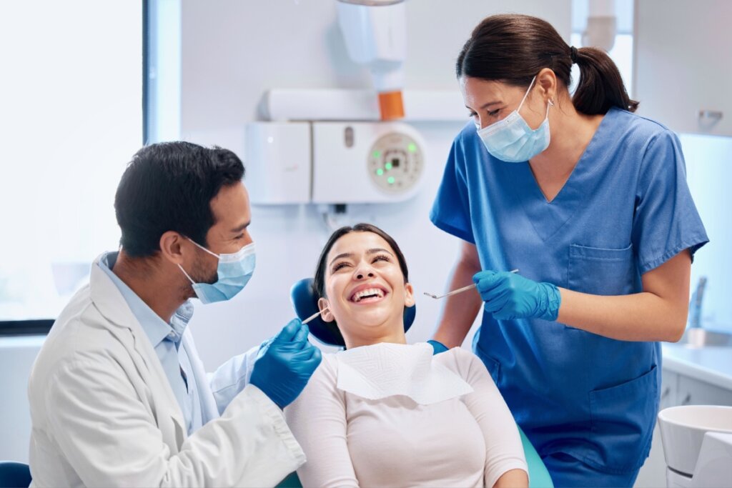Dois dentistas com jalecos brancos atendendo uma paciente sentada na cadeira odontológica