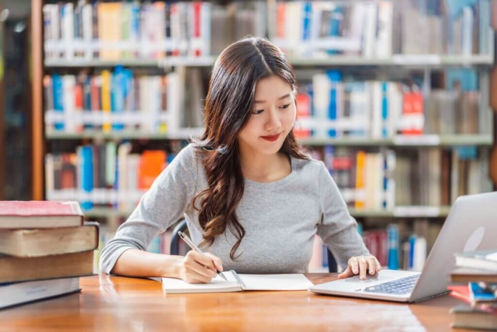 menina jovem de blusa cinza sentada em biblioteca, escrevendo em caderno e mexendo em notebook