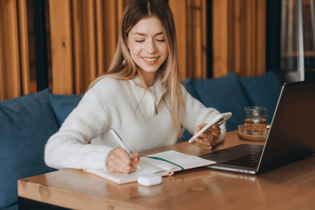 Estudante com cabelo longo, liso e loiro, usando blusa de lã branca fazendo anotações em agenda com celular na mão e notebook na mesa