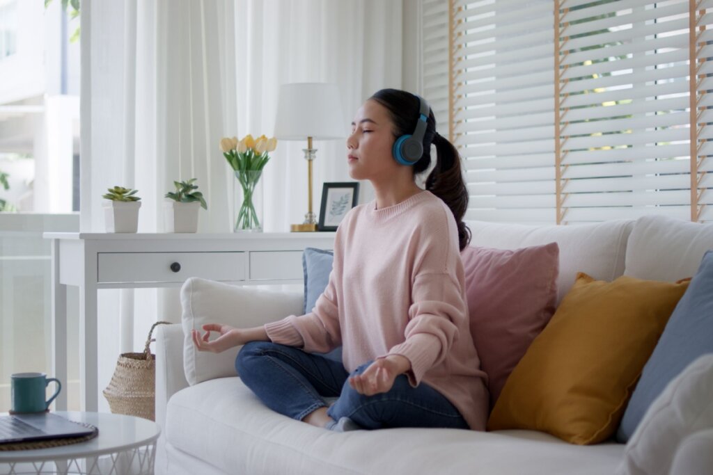 menina com headfone e blusa rosa sentada em sofá claro e meditando com as pernas cruzadas