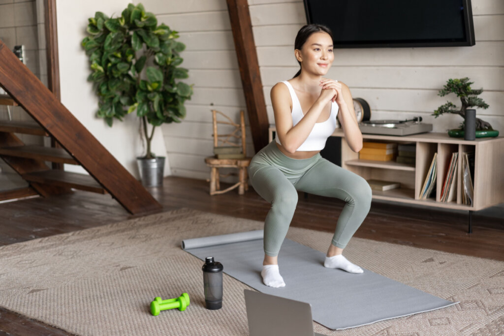 Uma mulher jovem fazendo agachamento sobre um colchonete cinza na sala de estar de sua casa. Ela veste um top branco e calça legging verde-clara. À sua frente, no chão, há um notebook, dois halteres verdes pequenos e uma coqueteleira de água.