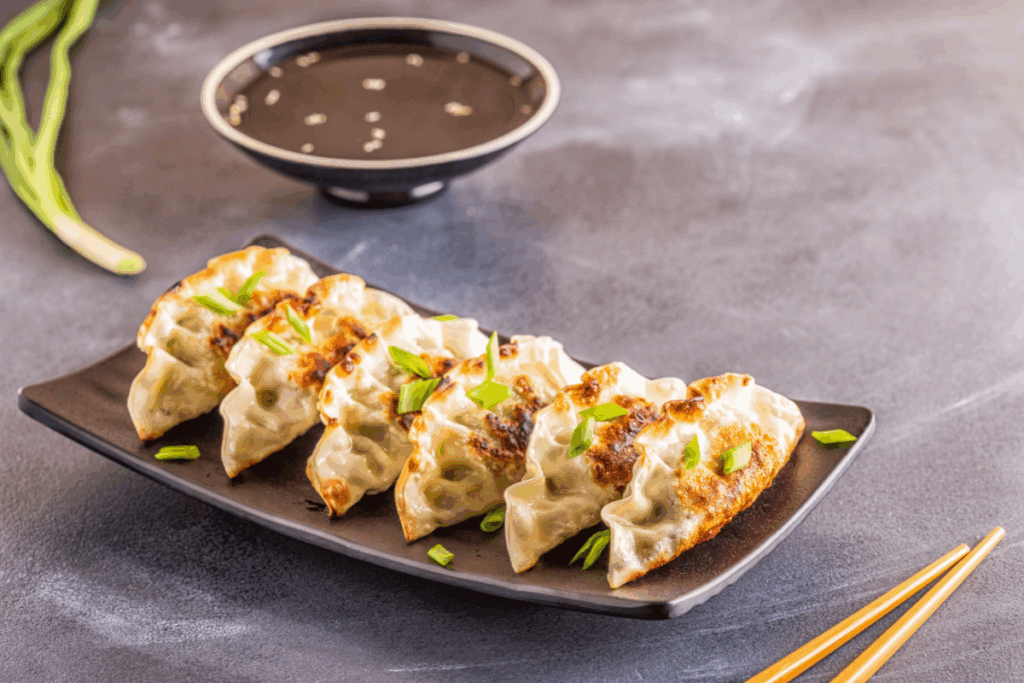 A imagem mostra gyozas grelhados dispostos em uma travessa preta, decorados com cebolinha picada e acompanhados de molho para mergulhar