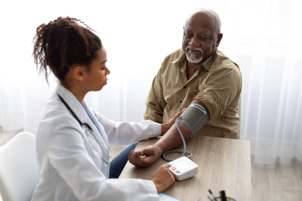 Uma foto simplificada mostrando uma médica jovem e negra de jaleco branco medindo a pressão arterial de um paciente idoso e negro sentado à sua frente, usando um esfigmomanômetro digital sobre a mesa.