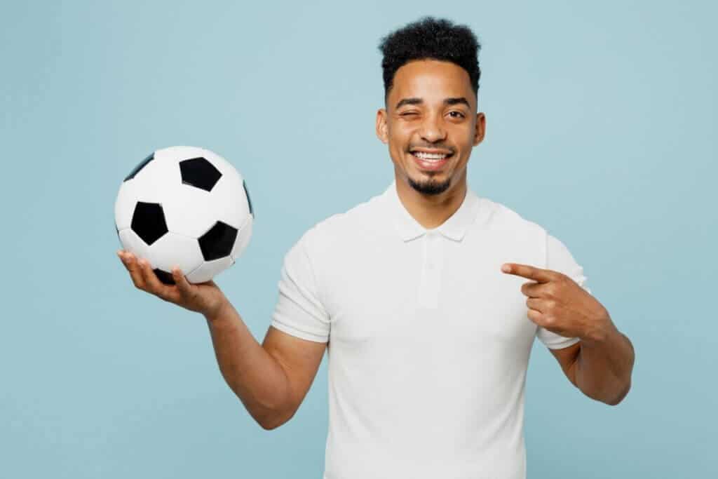 A imagem mostra um homem sorridente segurando uma bola de futebol com uma das mãos, enquanto aponta para ela com a outra. Ele veste uma camisa polo branca e aparece diante de um fundo azul-claro, transmitindo uma atmosfera leve e descontraída.