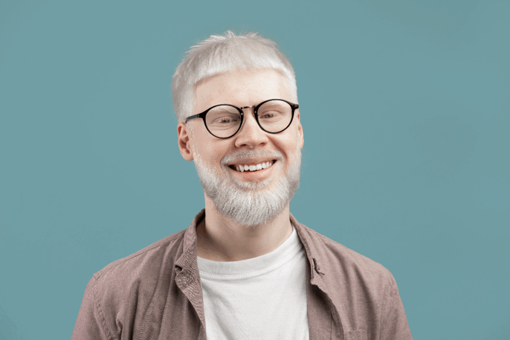 Homem albino com cabelo curto e barba, usando camiseta branca, camisa em tom de marrom claro e óculos preto