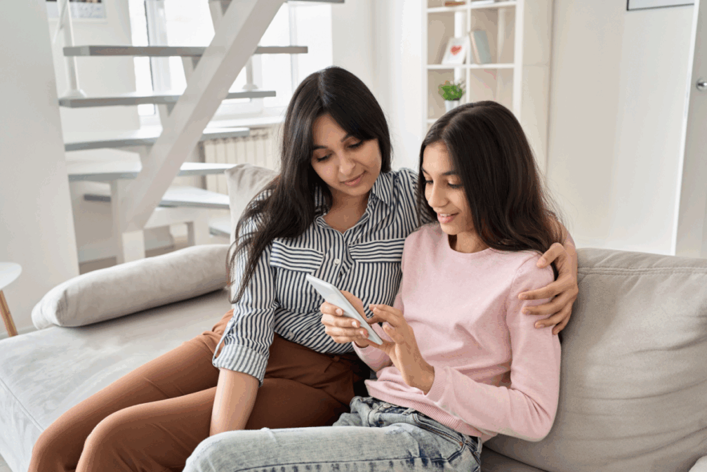 Mãe conversando com filha que está com o celular na mão, sentadas no sofá. A mulher tem o cabelo liso e solto, e está usando camisa cinza e branca listrada e calça marrom e a menina tem o cabelo liso, solto e está usando camisa de manga longa rosa e calça jeans