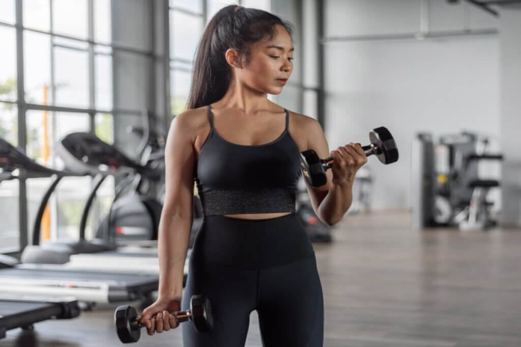 mulher de cabelo preso, regata e calça pretas, segurando halteres nas mãos e flexionando os braços