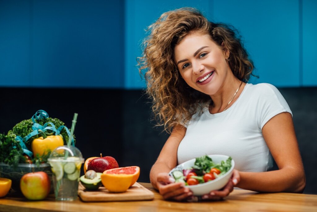 mulher de cabelo cacheado loiro, camiseta branca e segurando pote branco com salada. mesa ao lado com frutas e vegetais