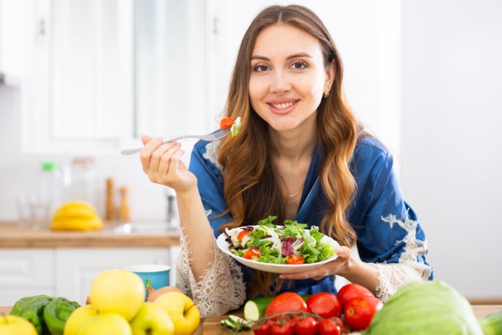 Mulher com cabelo castanho claro, ondulado solto, usando camisa jeans e comendo salada encostada na bancada da cozinha onde estão outros alimentos saudáveis