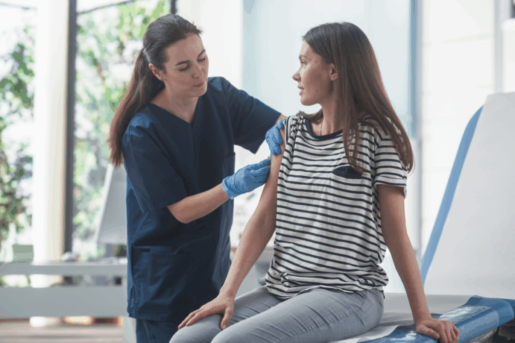 Enfermeira com cabelo longo, liso, preso usando uniforme de hospital azul escuro, luvas azuis aplicando vacina em mulher com o cabelo liso, solto, usando camiseta listrada preta e branca e calça jeans em sala de hospital