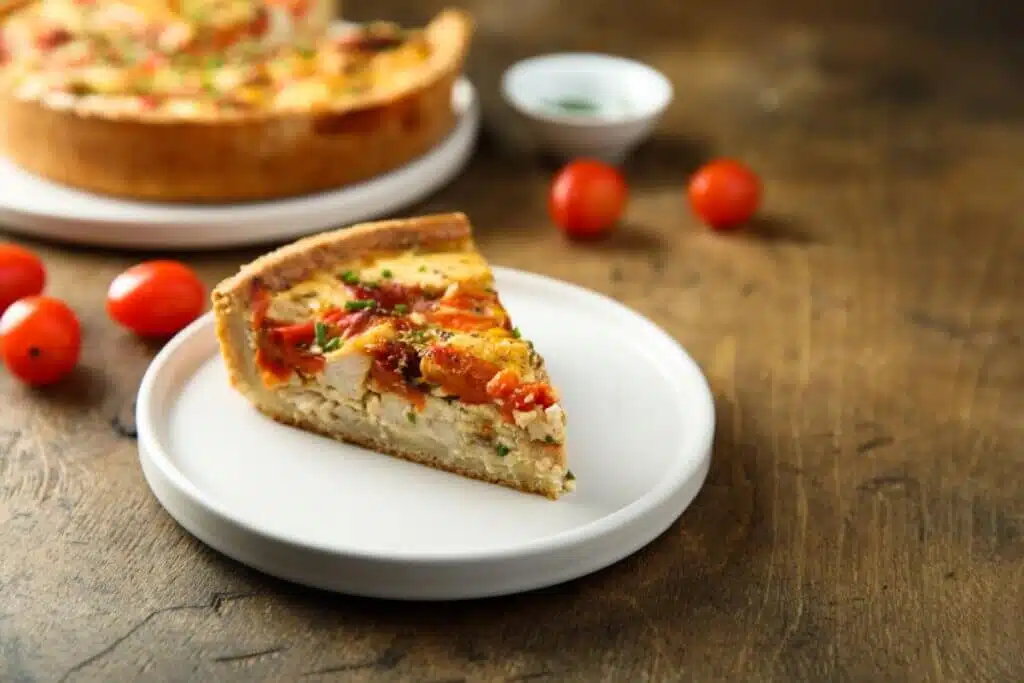 A imagem mostra uma fatia de quiche servida em um prato branco, com recheio aparente de frango desfiado e tomates-cereja assados. A massa é dourada e firme, enquanto o recheio tem textura cremosa, sugerindo uma combinação de ovos, queijo e temperos.