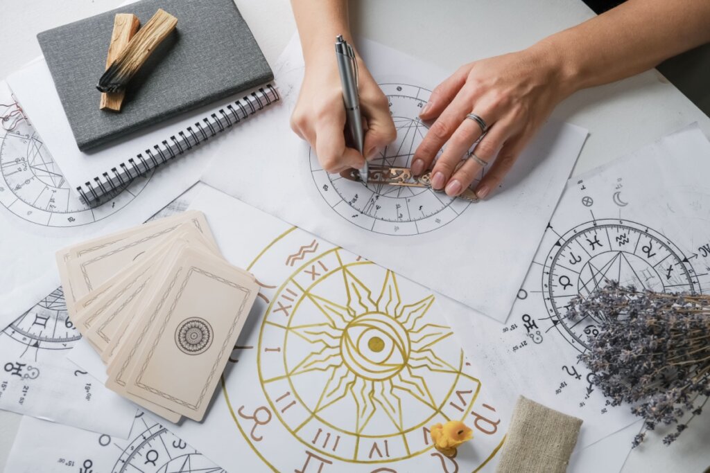 Mãos fazendo um mapa astral em um papel e rodeada por papéis com desenhos do mapa astral e cartas de tarô