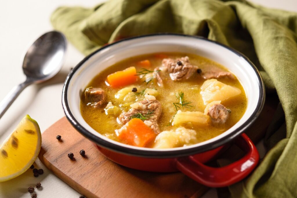 A imagem mostra uma sopa caseira servida em uma tigela vermelha, com pedaços de carne bovina, batata, cenoura e outros legumes cozidos em um caldo dourado e levemente encorpado
