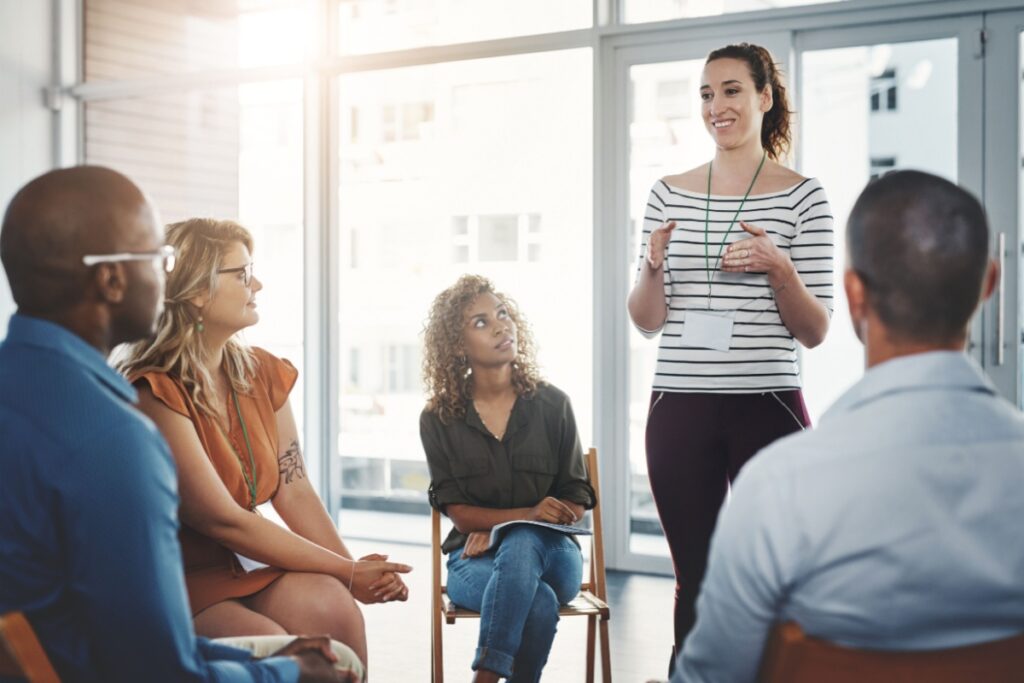 Mulher em pé falando para um grupo de homens e mulheres sentados