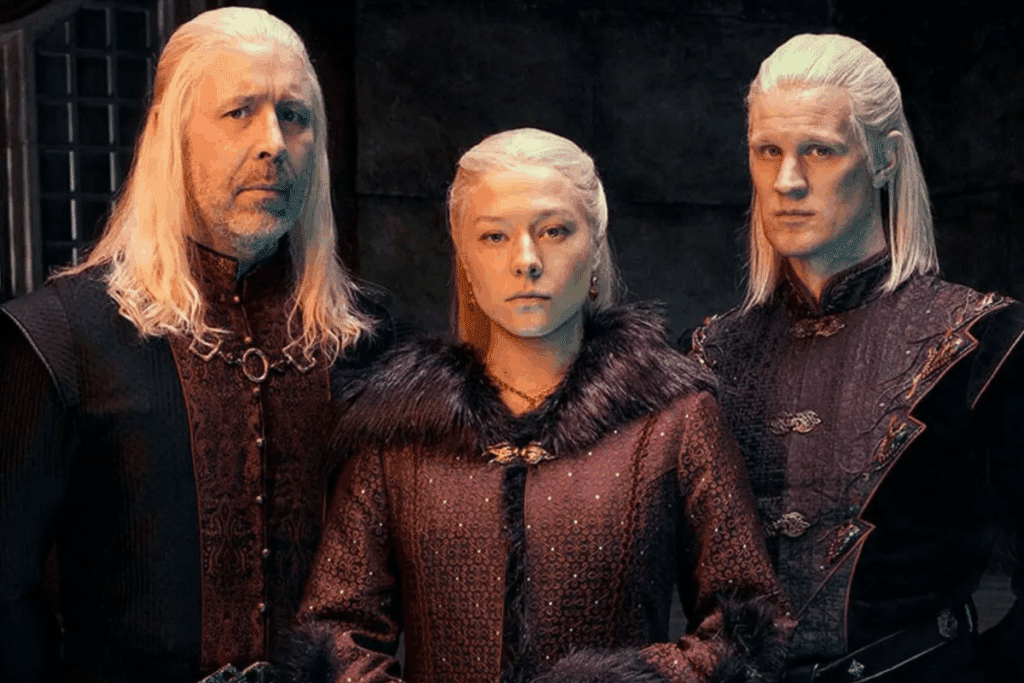 Três personagens com longos cabelos loiro-platinados posam lado a lado. No centro, uma mulher séria (Rhaenyra) veste trajes escuros com gola de pelos. Nas laterais, dois homens (Rei Viserys I e Príncipe Daemon Targaryen) usam roupas nobres em tons de preto e vinho.