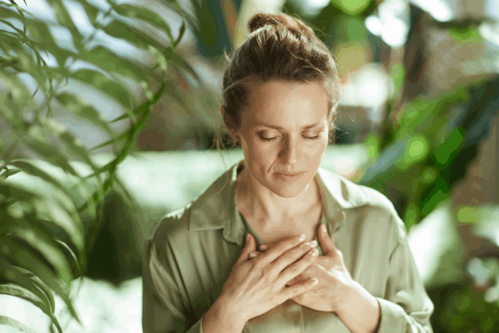 Mulher com os olhos fechados e expressão serena, segurando as duas mãos sobre o peito, cercada por plantas verdes ao fundo.