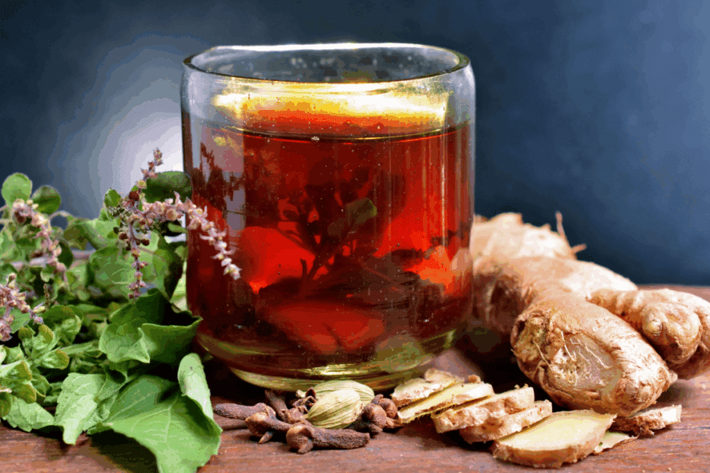 Copo de vidro transparente com chá escuro de alfavaca sobre superfície de madeira. Ao lado, ramos de alfavaca fresca com pequenas flores arroxeadas, pedaços e fatias de gengibre, cravos-da-índia e bagas de cardamomo.