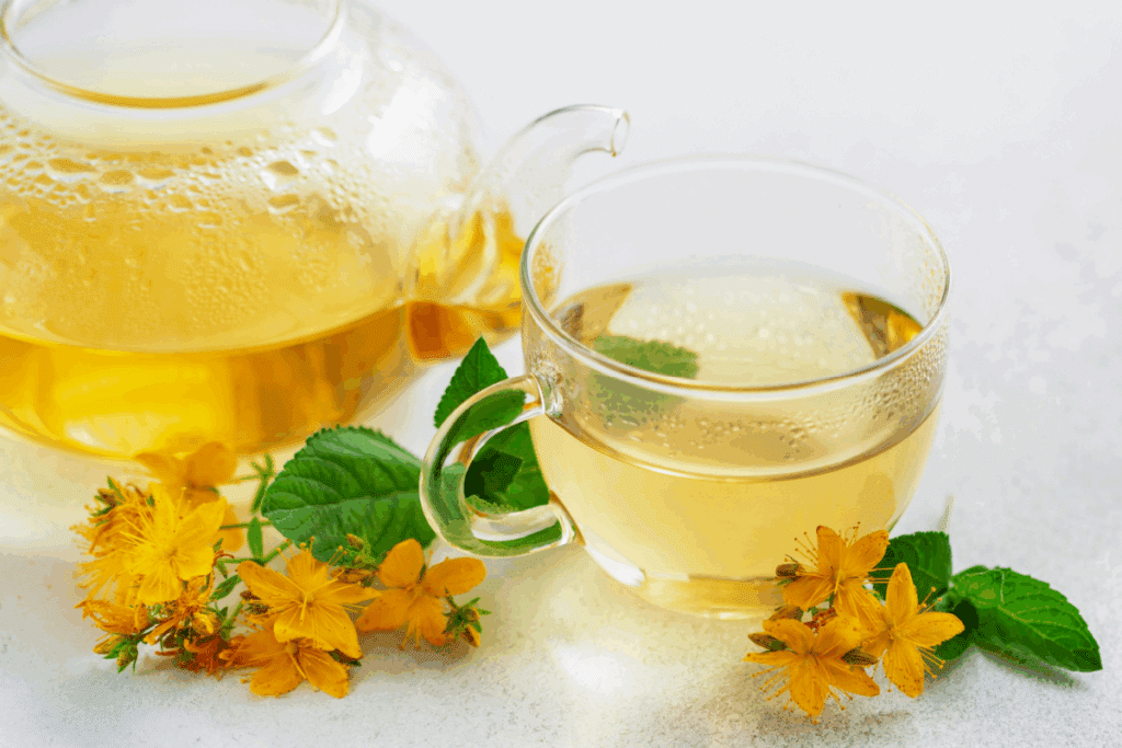 Xícara e bule de vidro transparente com chá amarelado, acompanhados de folhas verdes de hortelã e pequenas flores amarelas de erva-de-são-joão sobre uma superfície clara.
