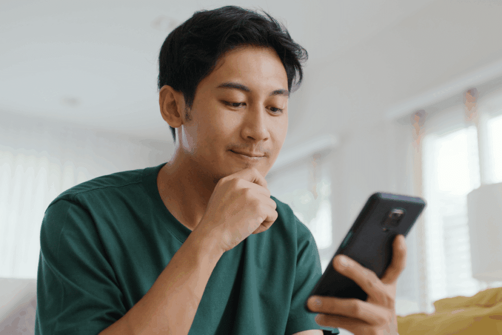 Homem jovem vestindo camiseta verde-escura olhando pensativo para a tela do celular que segura em uma das mãos, com o queixo apoiado na outra mão.
