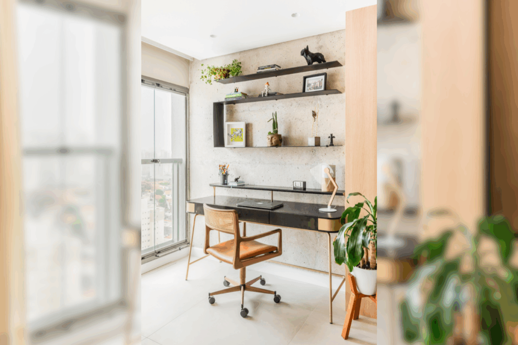 Espaço de home office montado em uma varanda fechada com vidro. O ambiente possui uma escrivaninha escura com pés dourados, uma cadeira de couro caramelo, prateleiras pretas na parede de concreto aparente e um vaso de planta no chão.