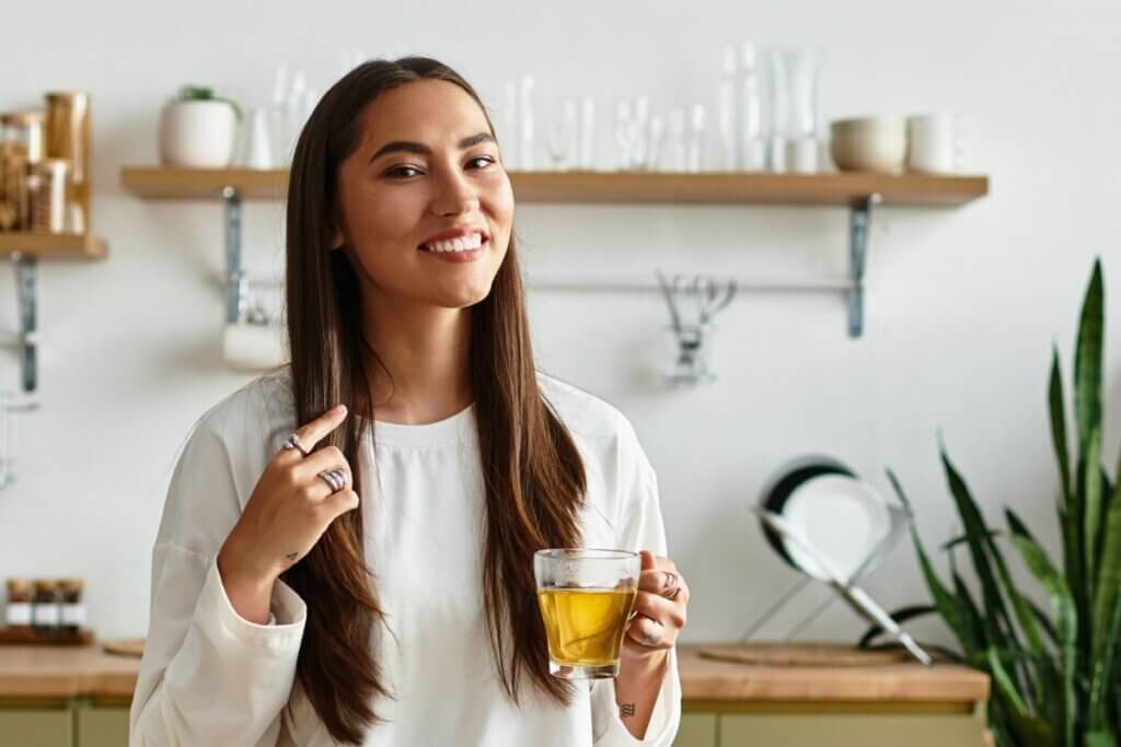 Uma jovem sorridente de cabelos longos e lisos segura uma caneca de vidro com chá claro em uma cozinha. Ela veste blusa branca e, ao fundo, aparecem prateleiras com utensílios e uma planta.