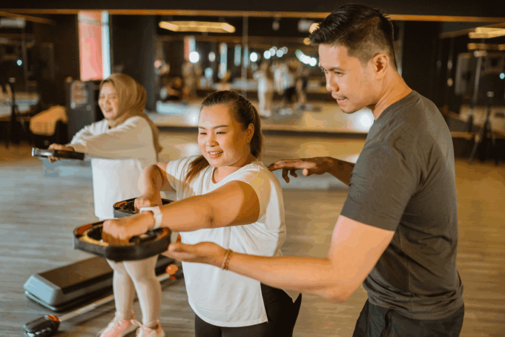 Um personal trainer orienta uma mulher que segura um peso de disco durante um treino em uma academia iluminada. Ao fundo, outra mulher também se exercita.