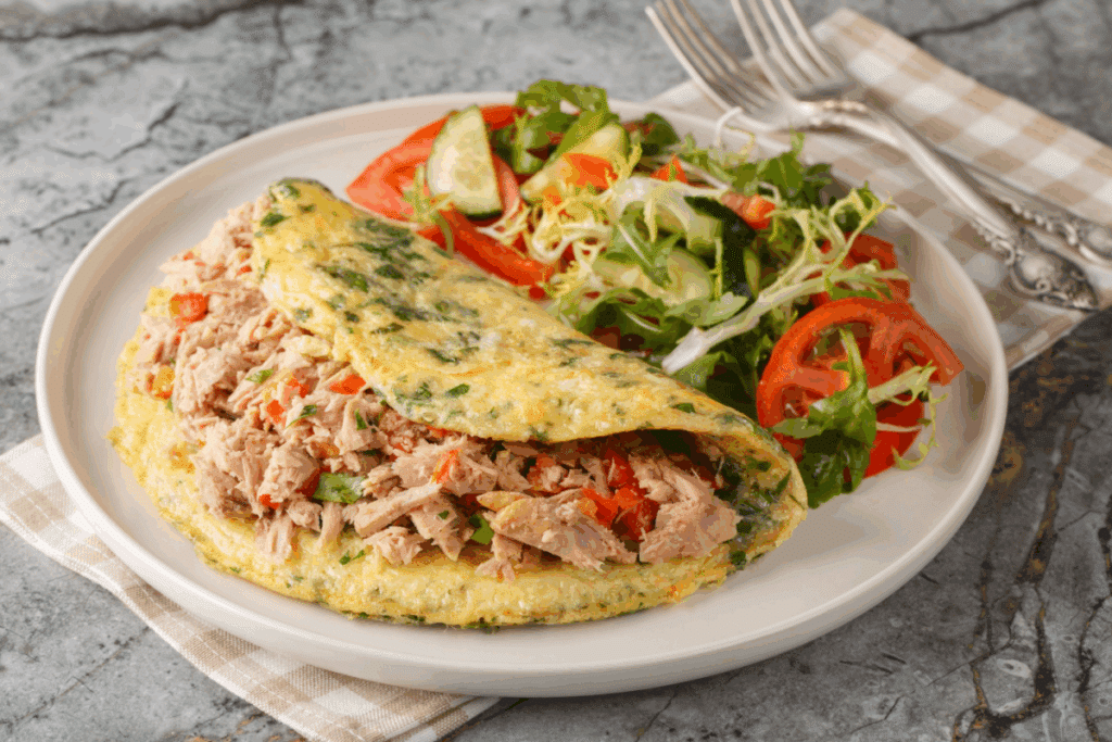 Uma omelete dobrada e recheada com atum desfiado e tomate, servida em um prato branco ao lado de uma salada verde mista com tomate e pepino.