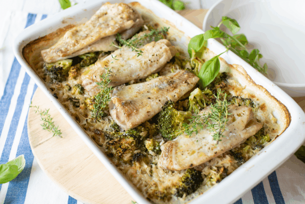 Travessa de cerâmica branca com filés de peixe assados acompanhados de brócolis e ervas frescas, como tomilho. A travessa está sobre uma tábua de madeira em cima de um pano listrado de azul e branco.