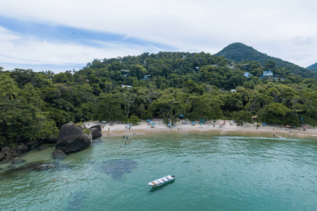 Vista aérea de uma praia cercada por mata verde densa. No mar verde-água, um pequeno barco a motor flutua no centro. À esquerda, grandes rochas escuras dividem a água, e na faixa de areia clara há guarda-sóis coloridos e pessoas aproveitando o dia.