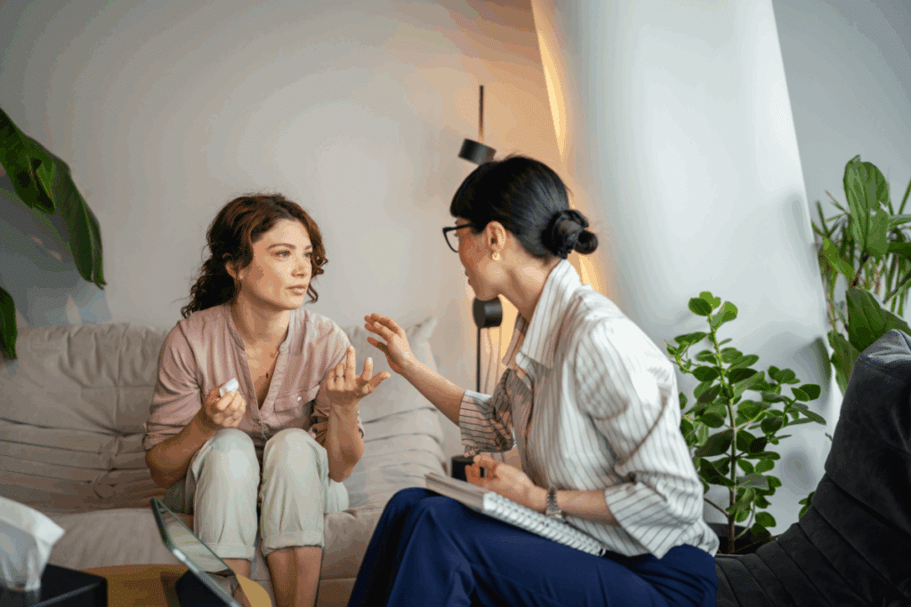 Foto em ambiente interno de uma sessão de terapia. À esquerda, uma mulher de cabelos cacheados e camisa rosa está sentada em um sofá claro, gesticulando com as mãos enquanto conversa. À direita, vista de perfil, a terapeuta — uma mulher de cabelos escuros presos em um coque, óculos e camisa listrada — segura um bloco de anotações no colo e estende levemente a mão em sinal de escuta empática. Ao fundo, há plantas verdes e iluminação suave.