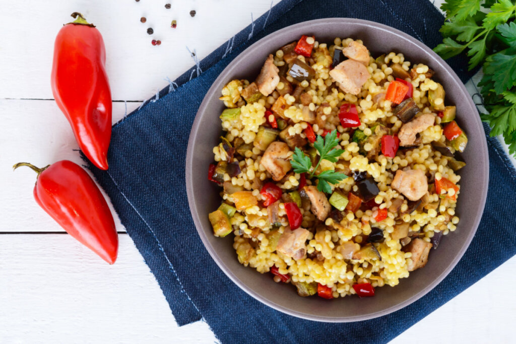 recipiente cinza com salada de cuscuz marroquino com frango, pimentão e berinjela. ao lado, dois pimentões vermelhos