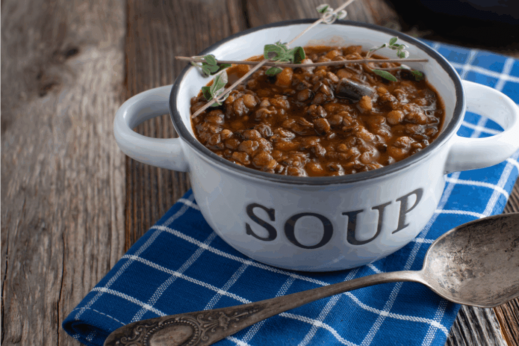 Uma tigela de cerâmica branca com alças e a palavra "SOUP" escrita em letras pretas, cheia de sopa densa de lentilhas com vegetais. O prato está decorado com ramos de ervas frescas no topo e posicionado sobre um pano xadrez azul e branco em uma mesa de madeira rústica, ao lado de uma colher antiga.
