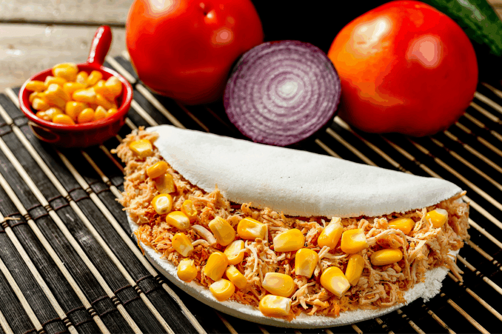 Tapioca dobrada ao meio recheada com frango desfiado e grãos de milho verde. Ao fundo, estão dispostos dois tomates, metade de uma cebola roxa e uma pequena colher de molho com milho.