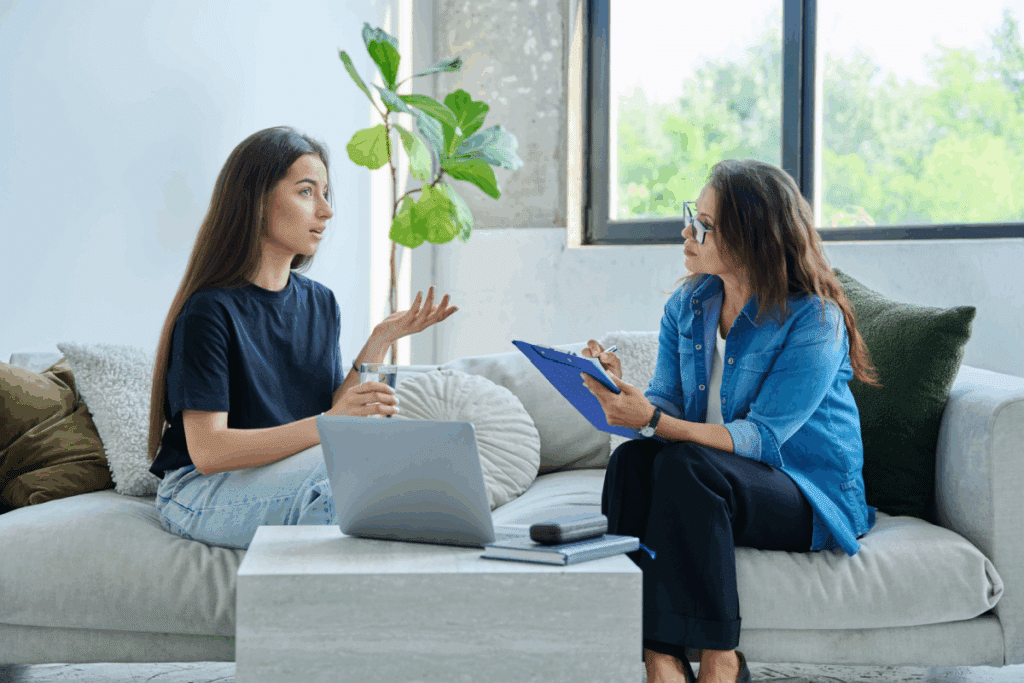 Duas mulheres sentadas num sofá claro durante uma sessão de terapia num consultório iluminado. À esquerda, uma jovem de camiseta preta e calça jeans fala gesticulando com uma mão, enquanto segura um copo de água na outra. À direita, a terapeuta de óculos, camisa azul e calças escuras, escuta-a com atenção enquanto toma notas numa prancheta azul. Sobre a mesa de centro à frente delas, há um computador portátil fechado e um caderno.