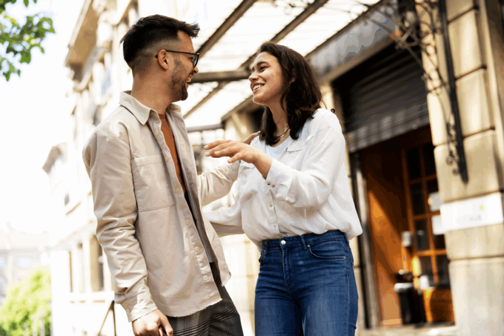 Um casal jovem e sorridente conversa de forma animada e descontraída em uma calçada urbana, com um edifício clássico ao fundo. A interação demonstra proximidade e alegria enquanto interagem ao ar livre.