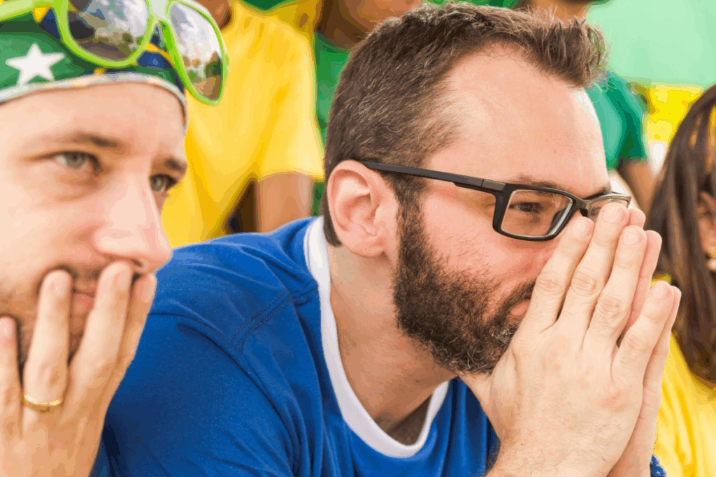 Homem, usando óculos de grau e camisa azul, inclinado com as duas mãos no rosto e assistindo jogo de futebol