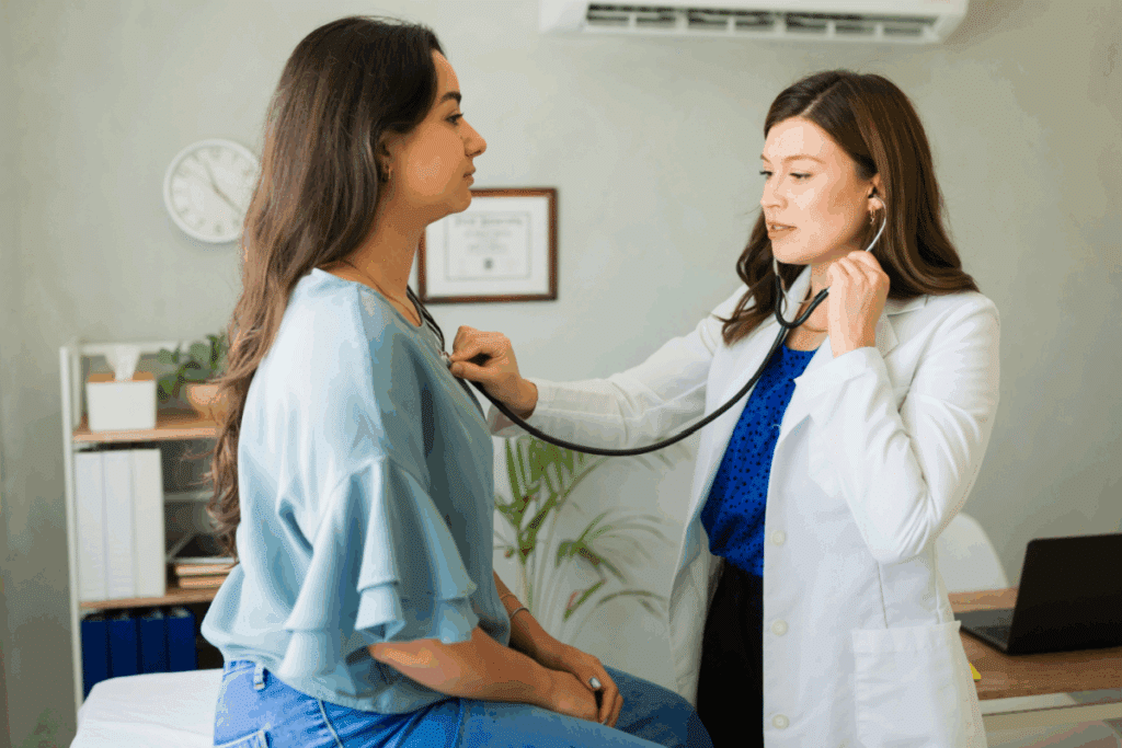 Médica com cabelo curto, usando blusa azul-escura, calça preta ouvindo pulmão de paciente com cabelo longo, usando camisa azul com babados e calça jeans sentada em maca de consultório