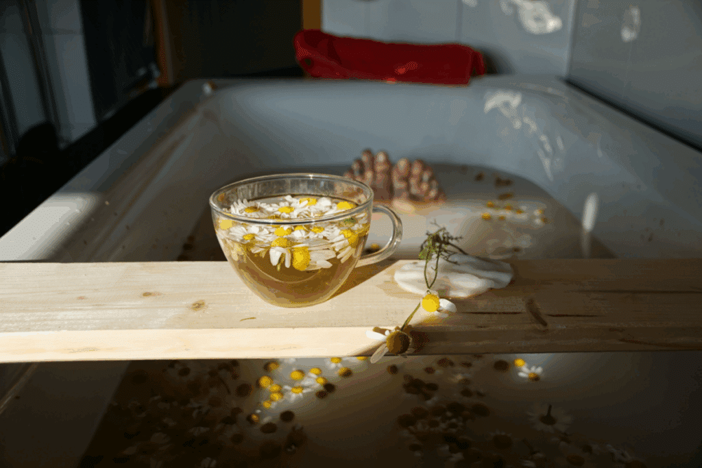 Uma banheira branca cheia de água de aspecto leitoso, com diversas flores de camomila flutuando na superfície. Ao fundo, os pés de uma pessoa que está tomando banho e uma tábua de madeira atravessando a banheira com uma xícara de chá de camomila 