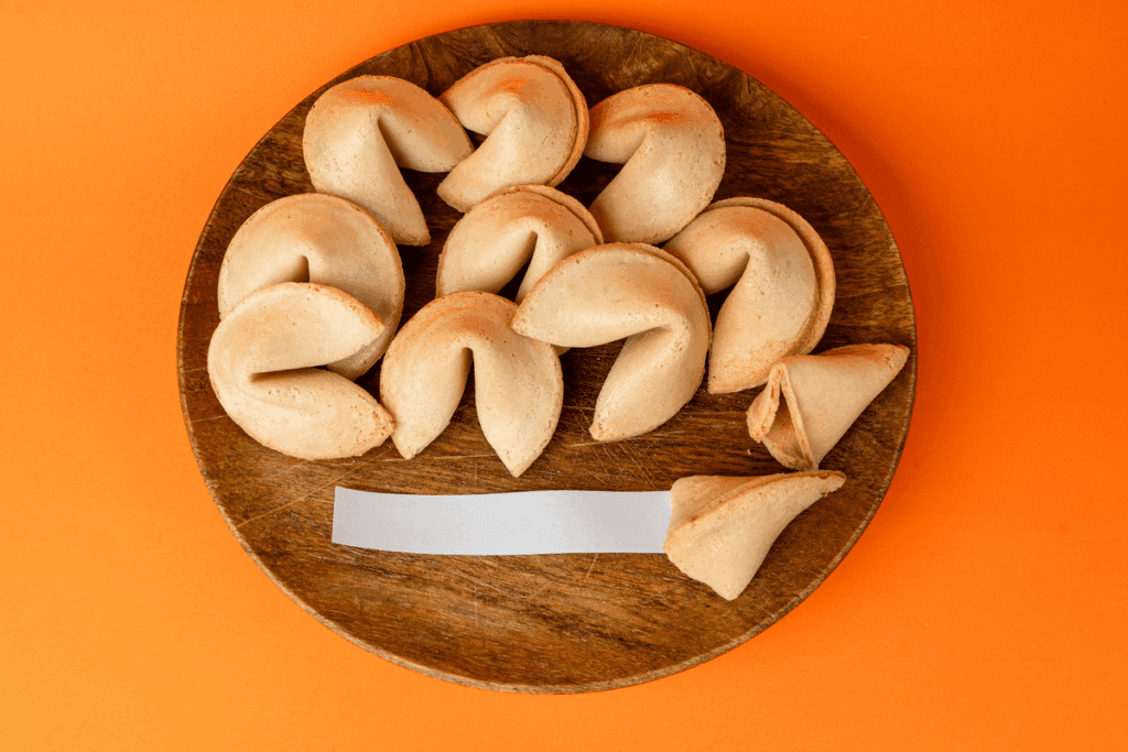 Vários biscoitos da sorte sobre um prato de madeira redondo, com um papel branco em branco à frente, em fundo laranja