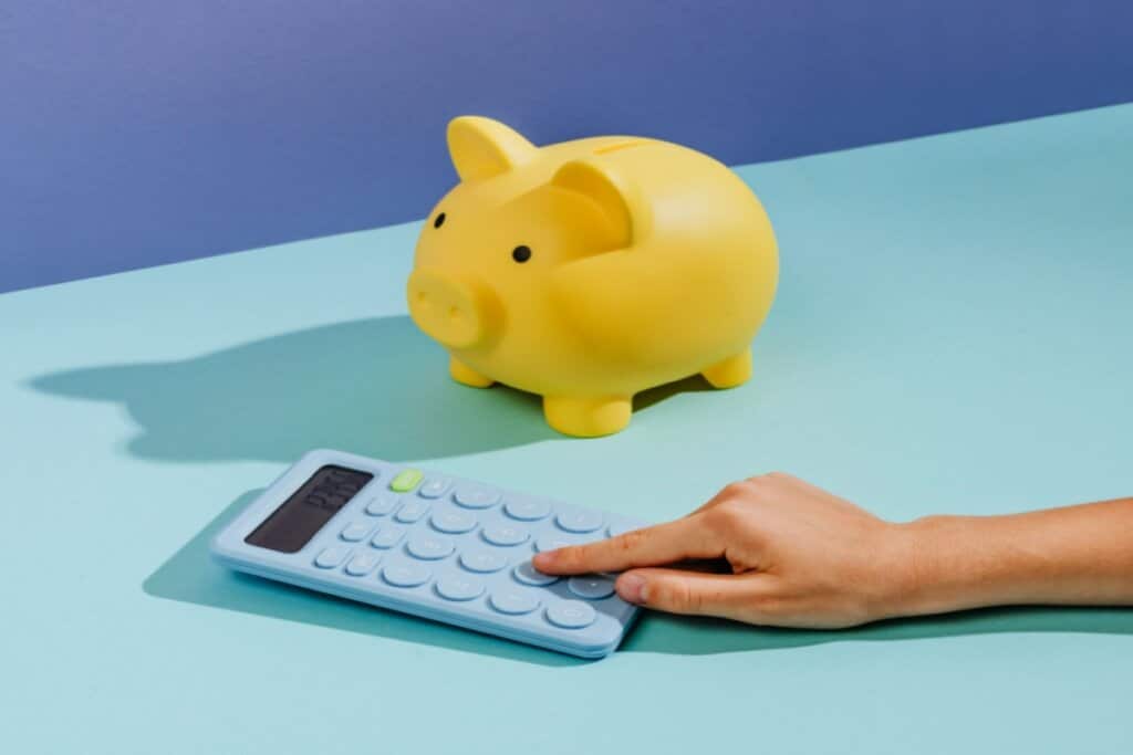 mão sobre calculadora azul ao lado de cofrinho amarelo em fundo azul-claro e escuro