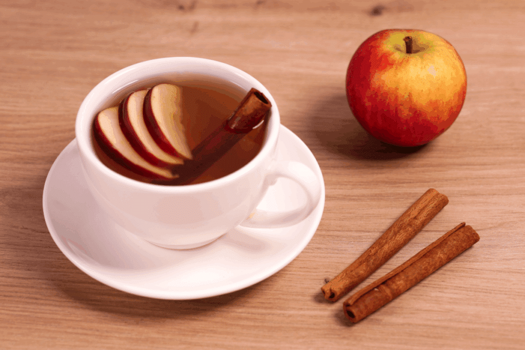 Xícara branca com chá de canela com maçã em mesa de madeira. Ao lado, duas canelas em pau e uma maçã