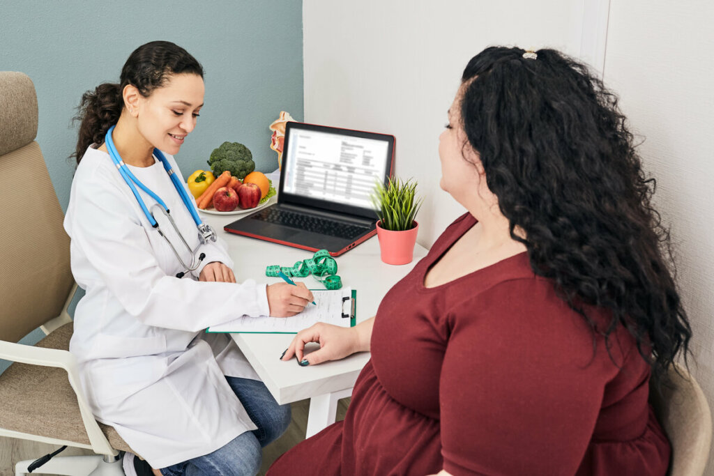 mulher de blusa roxa sentada ao lado de médica, que usa jaleco branco e escreve em papel. na mesa, há notebook e prato com frutas e vegetais