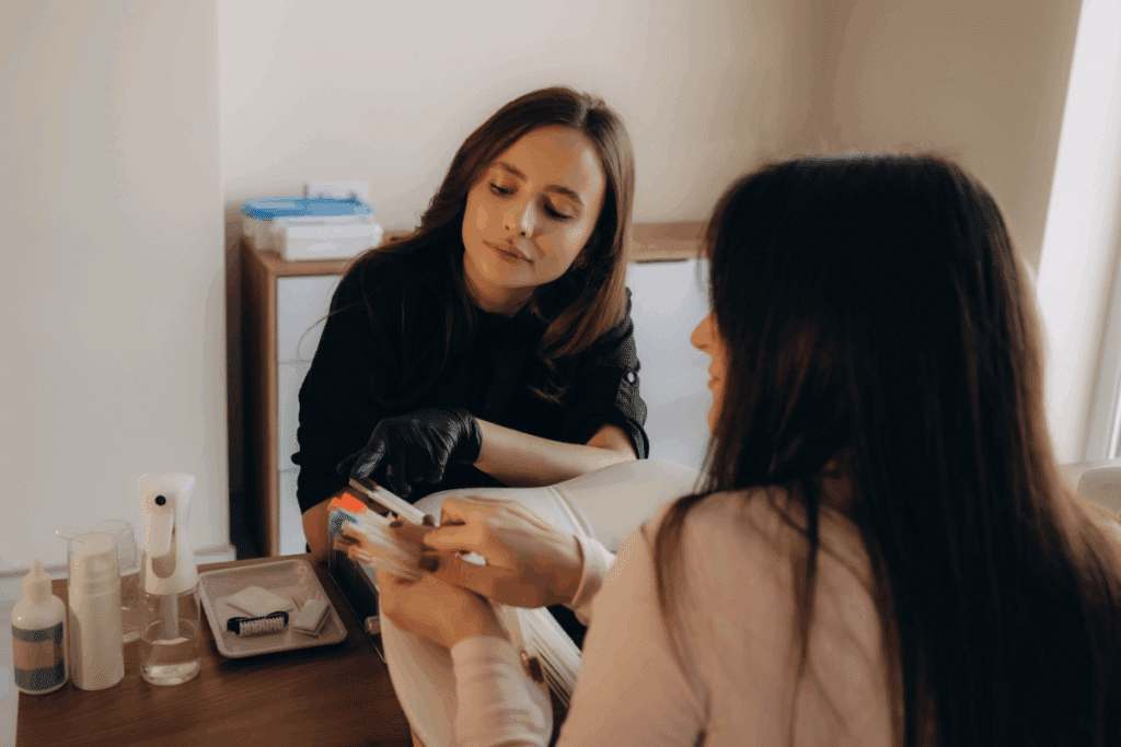 Mulher mostrando cor em paleta para uma manicure, ambas sentadas em lados opostos de uma mesa