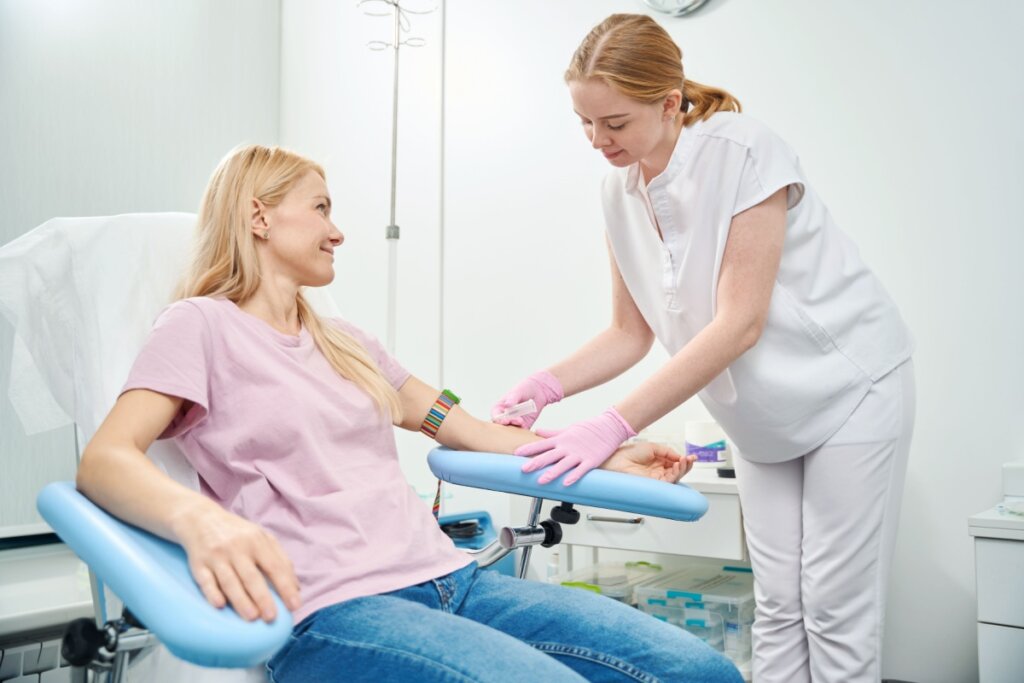 mulher de camiseta rosa sentada em cadeira azul e enfermeira ao lado, em pé, retirando sangue do braço dela