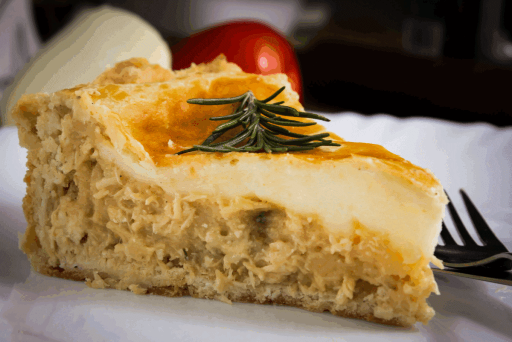 A imagem mostra uma fatia generosa de torta de frango servida em um prato branco. A massa dourada e levemente gratinada cobre um recheio cremoso de frango desfiado, no qual é possível notar pedaços de cogumelos e temperos distribuídos pela mistura.