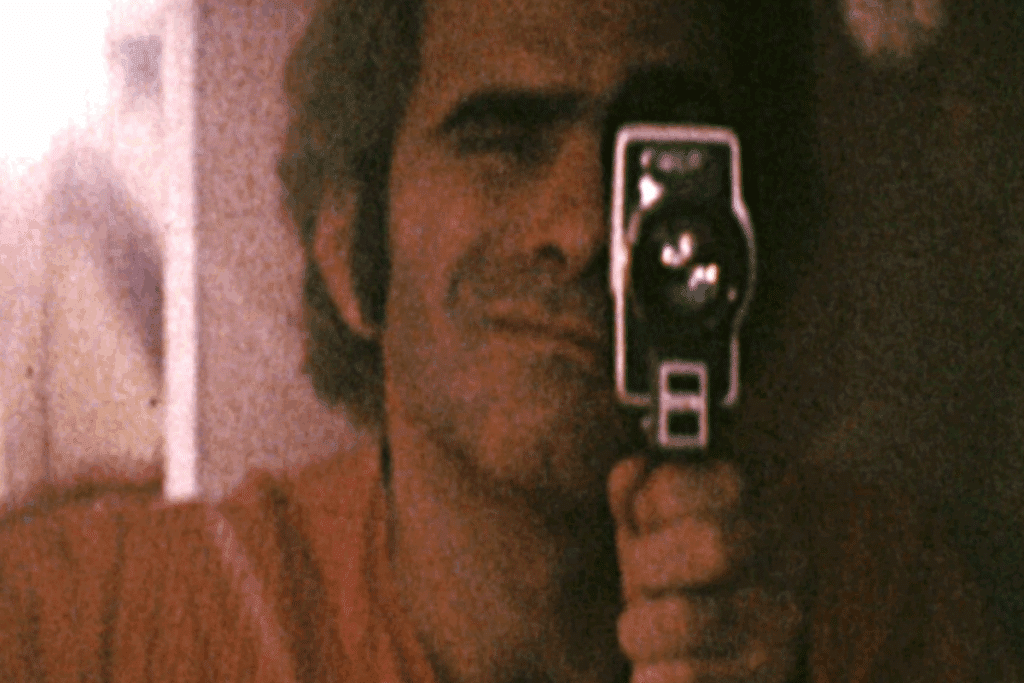 Imagem de um homem com o cabelo curto, usando camiseta laranja e com máquina antiga de filme perto dos olhos